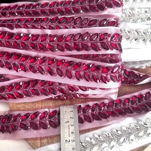 May include: Two rolls of beaded trim with a leaf design. One roll is pink with burgundy colored beads and the other is white with silver beads. A ruler is included for scale.