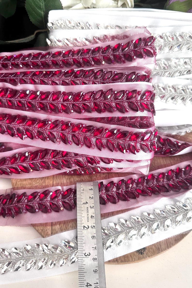 May include: Two rolls of beaded trim with a leaf design. One roll is pink with burgundy colored beads and the other is white with silver beads. A ruler is included for scale.