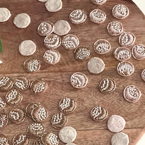 May include: Assortment of round, embroidered fabric embellishments in gold and cream colors. Some embellishments are beaded with white pearls.