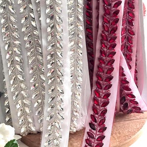 Puede incluir: Dos rollos de ribete con un diseño de hojas. Un rollo es blanco con pedrería transparente y el otro es rosa con pedrería roja.