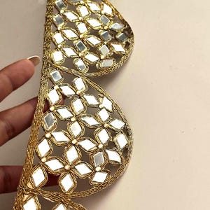 Op de afbeelding: Een decoratieve gouden bies met een geschulpt randontwerp. De bies is geconstrueerd met metallic gouden draadborduurwerk en ingelegde ruitvormige spiegels die in bloemachtige patronen over de geschulpte secties zijn gerangschikt.