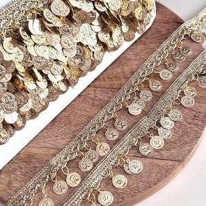 Könnte beinhalten: Goldmünzenbesatz mit aufwendigen Details. Der Besatz zeigt Reihen kleiner, baumelnder Münzen und einen dekorativen Rand. Die Münzen haben einen Vintage-Look und verleihen einen Hauch von Eleganz. Ideal zum Verzieren von Kleidung, Accessoires oder Bastelprojekten.