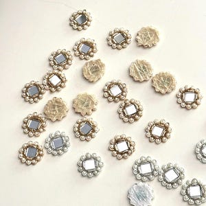 May include: A collection of decorative embellishments. The embellishments feature a square mirror center surrounded by small faux pearls. Some have a gold-colored border, while others have a silver-colored border. Several have a beige, textured fabric backing.
