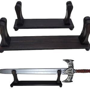 May include: Dark stained wooden sword display stands. The stands have a rectangular base with two curved supports. A silver and brown sword is displayed on one of the stands. The sword has an ornate hilt.