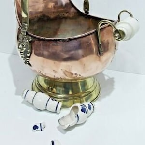 May include: A vintage copper and brass coal scuttle with a white porcelain handle and a base. The scuttle is sitting on a white surface and there are several broken pieces of white porcelain with blue floral designs scattered around it.