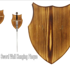 May include: A wooden shield-shaped plaque with two hooks for hanging swords. The plaque is made of light brown wood with a natural wood grain finish. "Sword Wall Hanging Plaque" is printed in black text at the bottom of the plaque.