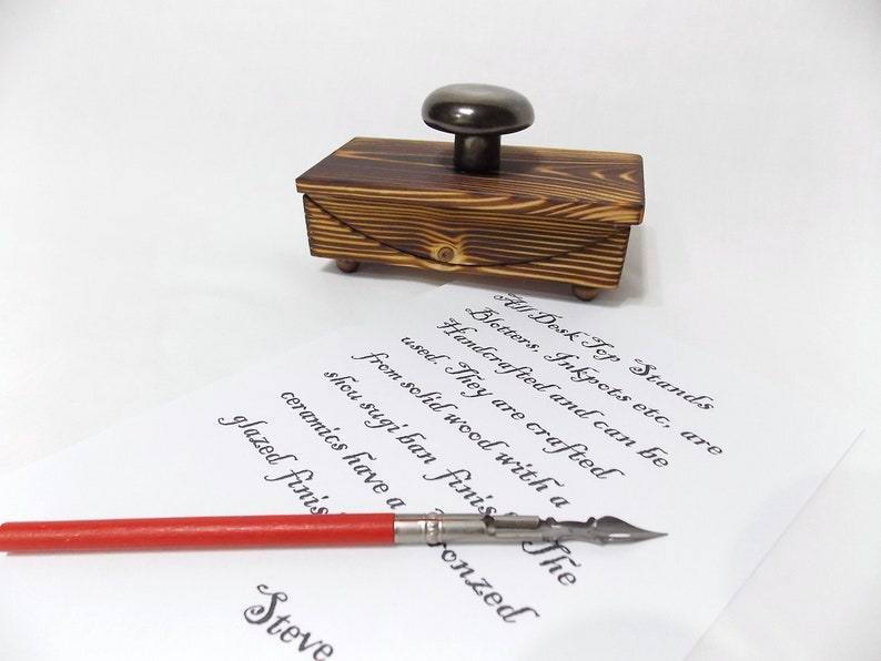 K&ouml;nnte beinhalten: Ein Tintenfass aus Holz mit einem Metalldeckel und einer Kalligraphie-Feder mit rotem Griff. Das Tintenfass ist handgefertigt aus massivem Holz mit einer Shou Sugi Ban-Oberfl&auml;che. Der Text auf dem Papier lautet: "All Desk Top Stands, Bletters, Inkpots etc, are Handcrafted and can be used. They are crafted from solid wood with a shou sugi ban finish. The ceramics have a glazed finish."