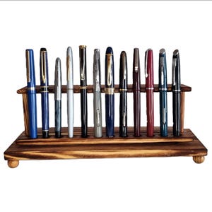 Op de afbeelding: Een houten pennenstandaard met twaalf vulpennen. De pennen zijn in verschillende kleuren, waaronder blauw, zilver, zwart en bordeauxrood. De standaard is gemaakt van gebeitst hout en heeft een ontwerp met twee niveaus.