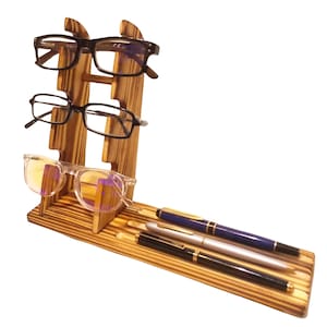 May include: A wooden eyeglasses and pen organiser. The organiser has three slots for glasses and a flat base with three pens. The wood has a natural finish. Three pairs of eyeglasses are displayed.