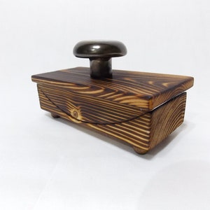 K&ouml;nnte beinhalten: Eine kleine Holzkiste mit einem Scharnierdeckel und einem Metallknopf oben. Die Kiste ist aus dunklem Holz mit einer Holzmaserung. Die Kiste steht auf einer wei&szlig;en Oberfl&auml;che.