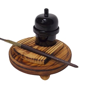 Puede incluir: Un tintero negro con tapa sobre un soporte de madera con una pequeña plataforma de madera y una pluma de madera apoyada en la plataforma.