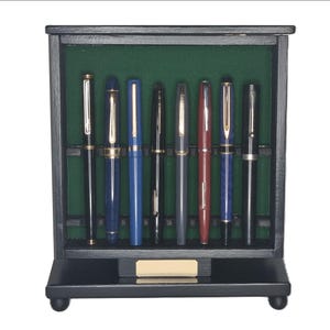 May include: A black wooden display case with a green felt interior holding eight fountain pens of various colours including black, blue, and burgundy. The case features a rectangular gold-coloured plaque at the base and stands on four small feet.