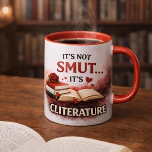 Op de afbeelding: Witte keramische mok met een rode binnenkant en oor, gevuld met donkere vloeistof en stoom. De mok heeft de tekst "IT'S NOT SMUT... IT'S CLITERATURE" met boek- en rozenillustraties. De mok staat op een houten oppervlak.
