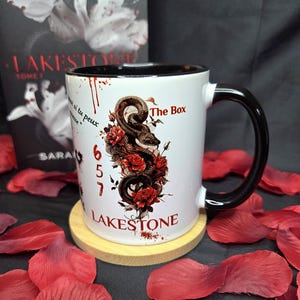Peut inclure: Mug en céramique blanche avec une anse et un bord noirs, orné d'un motif de serpent entrelacé de roses rouges. Le mug porte les inscriptions "LAKESTONE" et "The Box". Le mug est posé sur un dessous de verre en bois, entouré de pétales de roses rouges.