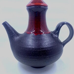 May include: A handmade ceramic teapot. The body is black, with a red-brown upper section and lid. It has a curved spout and a loop handle. The finish is glossy. The teapot is designed for serving tea.