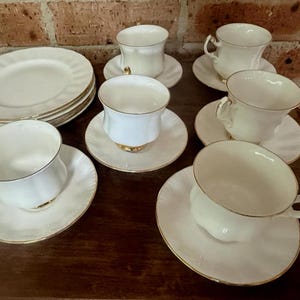 Pode incluir: Um conjunto de xícaras e pires brancos com detalhes dourados, juntamente com uma pilha de pratos combinando. As xícaras têm um design clássico, dispostas sobre uma superfície de madeira escura.