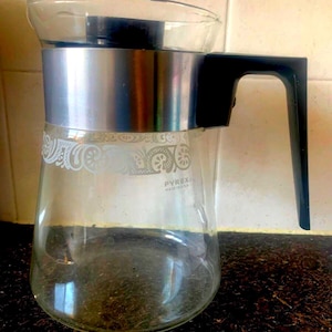 May include: A clear glass Pyrex jug with a silver band and black handle. The jug features a white floral pattern and the text "PYREX MADE IN USA".