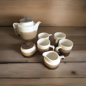 Puede incluir: Un juego de té de cerámica con tetera, azucarero, lechera y seis tazas. El juego es blanco, marrón y beige. La tetera tiene tapa y asa marrones. Las tazas son de dos tonos.