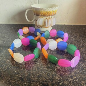 May include: A collection of colorful beaded jewelry, including two bracelets with large, oval beads in shades of pink, green, blue, orange, and white. A mug with gold trim holds several pairs of dangling earrings.