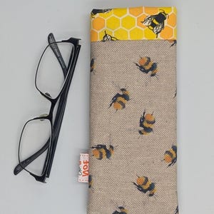 May include: A beige glasses case with a bumble bee pattern. The case has a yellow and orange honeycomb pattern on the top flap. The case is lying on a white surface next to a pair of black framed glasses.