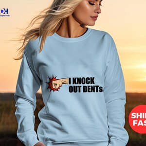 Puede incluir: Una sudadera de color azul claro con un gráfico de un puño golpeando las palabras "I knock out dents".