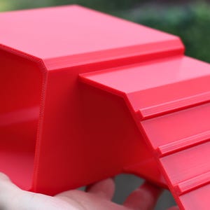 May include: A bright red, 3D-printed plastic ramp featuring a flat top platform and a series of horizontal ridges along the inclined surface to provide traction. The structure has a hollow underside and a smooth, matte finish throughout the design.