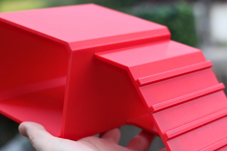 May include: A bright red, 3D-printed plastic ramp featuring a flat top platform and a series of horizontal ridges along the inclined surface to provide traction. The structure has a hollow underside and a smooth, matte finish throughout the design.