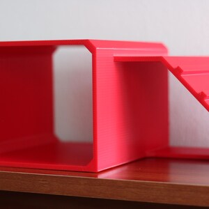 May include: A bright red, hollow, rectangular structure with an open front and back, featuring an attached ramp on the right side. The ramp has horizontal ridges along its surface, and the entire object is constructed from a solid, matte material.