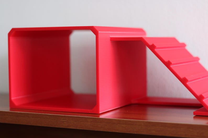 May include: A bright red, hollow, rectangular structure with an open front and back, featuring an attached ramp on the right side. The ramp has horizontal ridges along its surface, and the entire object is constructed from a solid, matte material.
