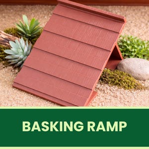 Könnte beinhalten: Eine rotbraune Sonnenrampe mit Lamellendesign, vor dem Hintergrund von Sand, Grün und einem Stein. Die abgewinkelte Rampe schafft einen schattigen Bereich. Der Text "BASKING RAMP" wird in einem grünen Banner unten angezeigt.