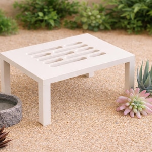 May include: A small, white, square pet stand with a slotted top. The stand is surrounded by decorative plants, a small water bowl, and a terracotta bowl with food. The scene is set on a bed of sand.