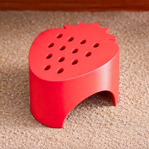 May include: A bright red, strawberry-shaped kitchen gadget. The top has a series of small holes, and the base is a curved, open design. The product is made of a solid material and sits on a textured surface.
