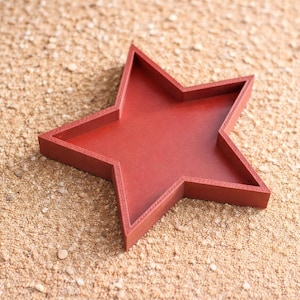 Può includere: Un vassoio rosso a forma di stella con bordi rialzati. Il vassoio è realizzato in un materiale solido e poggia su una superficie sabbiosa. La stella ha cinque punte e un interno piatto.