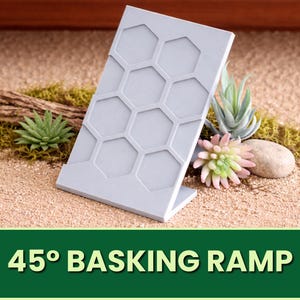 以下が含まれることがあります： ハニカム模様の、45度の角度のライトグレーの日光浴用ランプ。砂地の表面に、装飾用の植物と小さな石と一緒に展示されています。「45° BASKING RAMP」というテキストが緑色のバナーに表示されています。