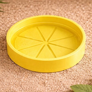 May include: A bright yellow, circular plant saucer with a segmented design, resembling a lemon slice. The saucer sits on a textured, sandy surface, with green leaves in the background. This decorative item is suitable for indoor or outdoor use.