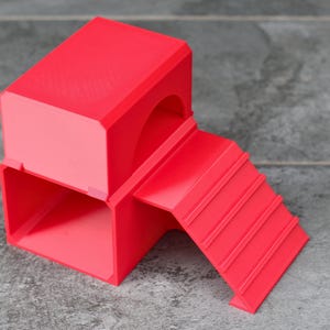May include: A bright red, 3D-printed structure featuring a hollow rectangular base with an open front, topped by a solid rectangular block with a curved archway entrance, and an attached ramp with horizontal ridges leading to the upper level.