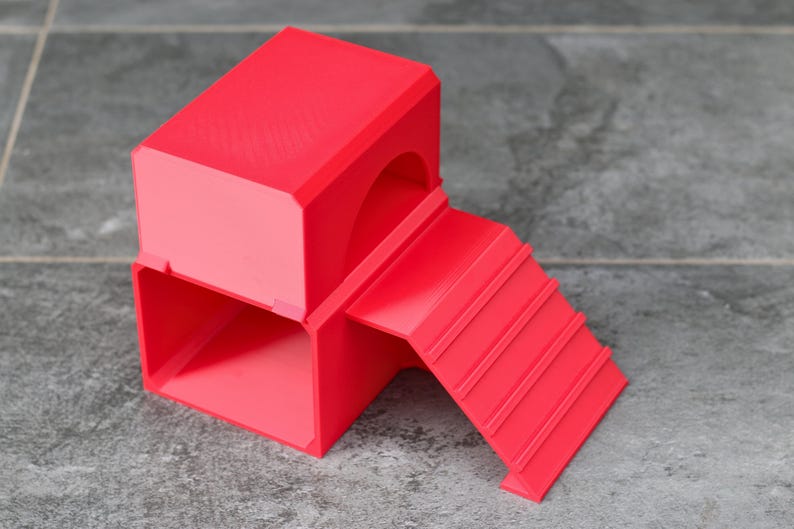 May include: A bright red, 3D-printed structure featuring a hollow rectangular base with an open front, topped by a solid rectangular block with a curved archway entrance, and an attached ramp with horizontal ridges leading to the upper level.