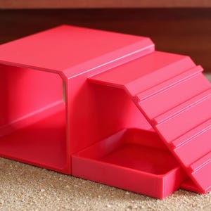 May include: A bright pink pet accessory featuring a hollow rectangular tunnel section connected to a ramp with horizontal ridges and a small square platform base. The structure is made of smooth, solid-colored plastic with a uniform matte finish.