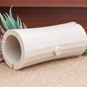 May include: A beige, cylindrical object with a circular opening and a small knob. The object has a segmented design and is placed on a sandy surface with a succulent plant in the background. The object is likely a decorative item.