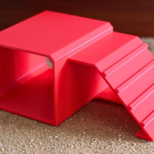May include: A bright red plastic pet hideout featuring a hollow rectangular tunnel structure with an attached ramp that has four horizontal ridges for traction. The object is constructed from a solid, smooth material with clean, angular edges.