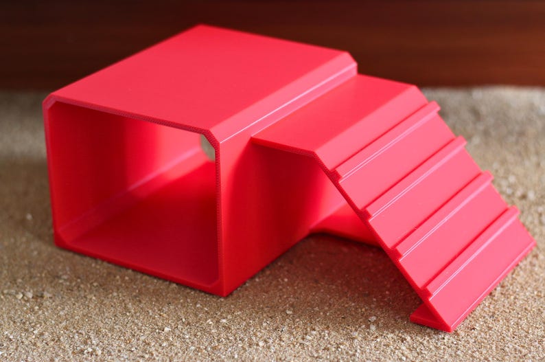 May include: A bright red plastic pet hideout featuring a hollow rectangular tunnel structure with an attached ramp that has four horizontal ridges for traction. The object is constructed from a solid, smooth material with clean, angular edges.