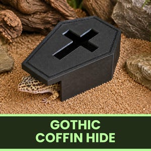 May include: A black coffin-shaped reptile hide with a cross-shaped cutout on the top lid. A spotted leopard gecko is partially visible underneath the structure. The object is constructed with a matte finish and features stitched detailing along the edges.