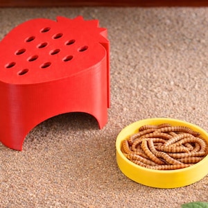 May include: A bright red, strawberry-shaped feeder with multiple holes, next to a yellow bowl filled with mealworms. The feeder has a curved base and is designed for small pets or reptiles.