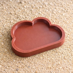 Puede incluir: Una bandeja en forma de nube de color terracota. La bandeja tiene un borde elevado y un interior liso. El producto está hecho de un material sólido y se encuentra sobre una superficie arenosa. Es un artículo decorativo.
