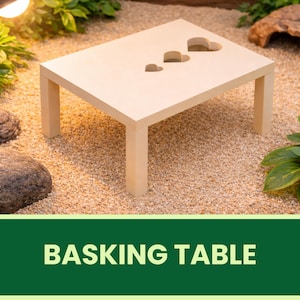 Könnte beinhalten: Ein helles Holz-Bade-Tisch mit drei herzförmigen Ausschnitten. Der Tisch hat vier quadratische Beine und steht auf einem Bett aus kleinen Kieselsteinen. Der Text "BASKING TABLE" wird in einem grünen Banner am unteren Bildrand angezeigt.