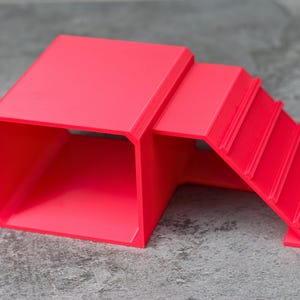 May include: A bright red, 3D-printed plastic structure featuring a hollow, rectangular tunnel section connected to a set of three stepped platforms. The object has a smooth, matte finish and a geometric design with clean, sharp edges throughout.