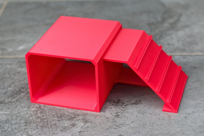 May include: A bright red, 3D-printed plastic structure featuring a hollow, rectangular tunnel section connected to a set of three stepped platforms. The object has a smooth, matte finish and a geometric design with clean, sharp edges throughout.
