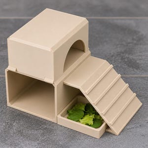 Peut inclure: Un abri pour animal en plastique beige composé d'une structure à deux niveaux avec une chambre inférieure rectangulaire, une entrée supérieure arquée et une rampe menant à un petit plateau carré contenant des légumes verts frais.