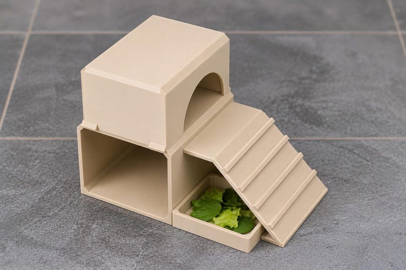 May include: A beige plastic pet hideout featuring a two-level structure with a rectangular lower chamber, an upper arched entrance, and an attached ramp leading to a small square feeding tray containing fresh green leafy vegetables.