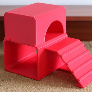 May include: A bright red plastic pet hideout featuring a two-level structure with a hollow square base and a top platform with an arched entrance. A set of stairs with four horizontal ridges is attached to the side, leading up to the top level.